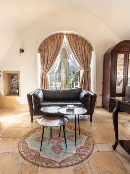 Elegant seating area with a leather sofa and a large arched window at Flora Valley Boutique Hotel, Ein Kerem, Jerusalem – a blend of luxury and warmth.